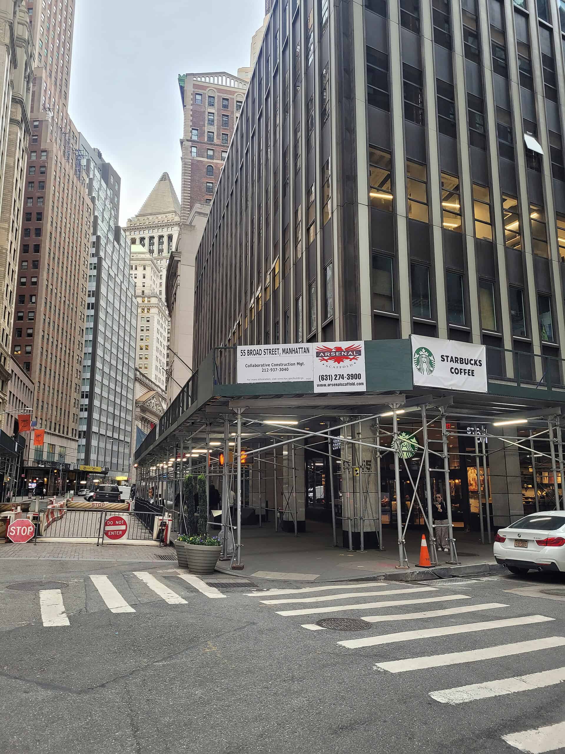 Base of 55 Broad Street in Manhattan during its office-to-residential conversion, with a sidewalk shed and contractor signage at the corner