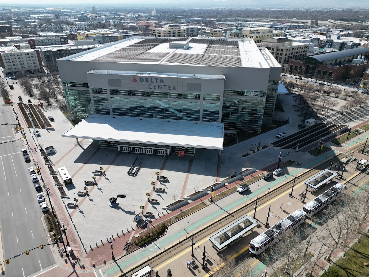 Aerial view of the Delta Center arena in downtown Salt Lake City, with its rooftop mechanical plant, entry canopy and a TRAX light rail train passing on the street below.