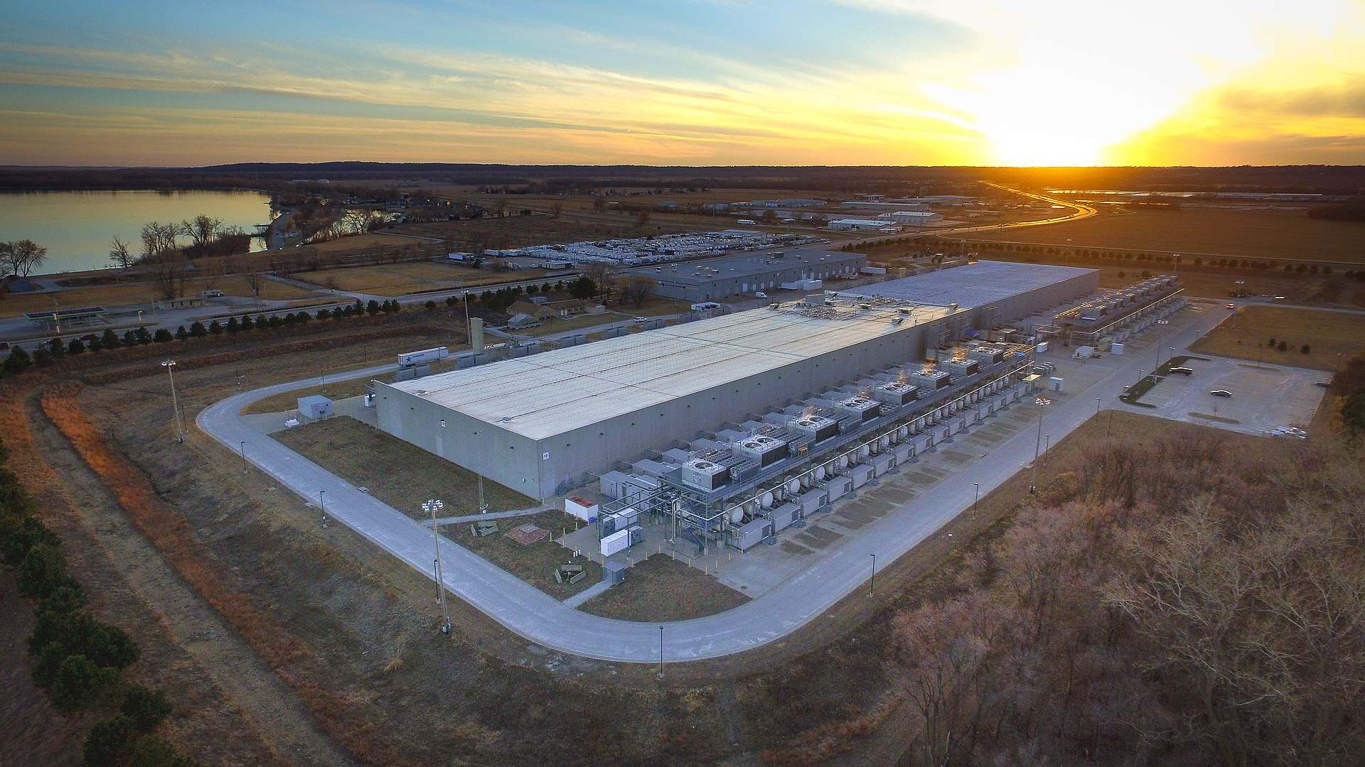 Aerial view at sunset of a long single-story data center building surrounded by parking and open farmland