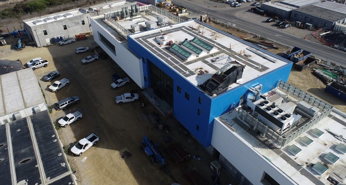 Aerial view of the North City Pure Water Facility in San Diego under construction, showing the white and blue process building, rooftop mechanical equipment, perimeter concrete structures and site laydown with Interstate 805 at the top of the frame.