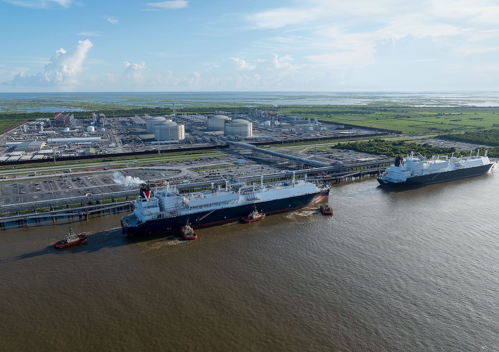 Aerial view of the Plaquemines LNG export terminal on the Mississippi River in Louisiana, with two LNG carriers berthed at the marine facility under tug escort, four white LNG storage tanks and the liquefaction trains behind the levee.