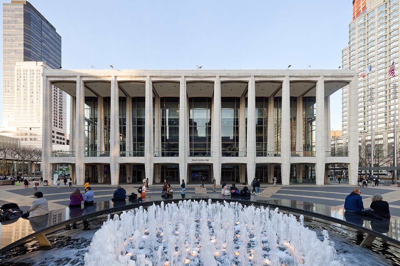 Projects David Geffen Hall's travertine colonnade and glass facade on Lincoln Center plaza, seen across the Revson Fountain in March 2024