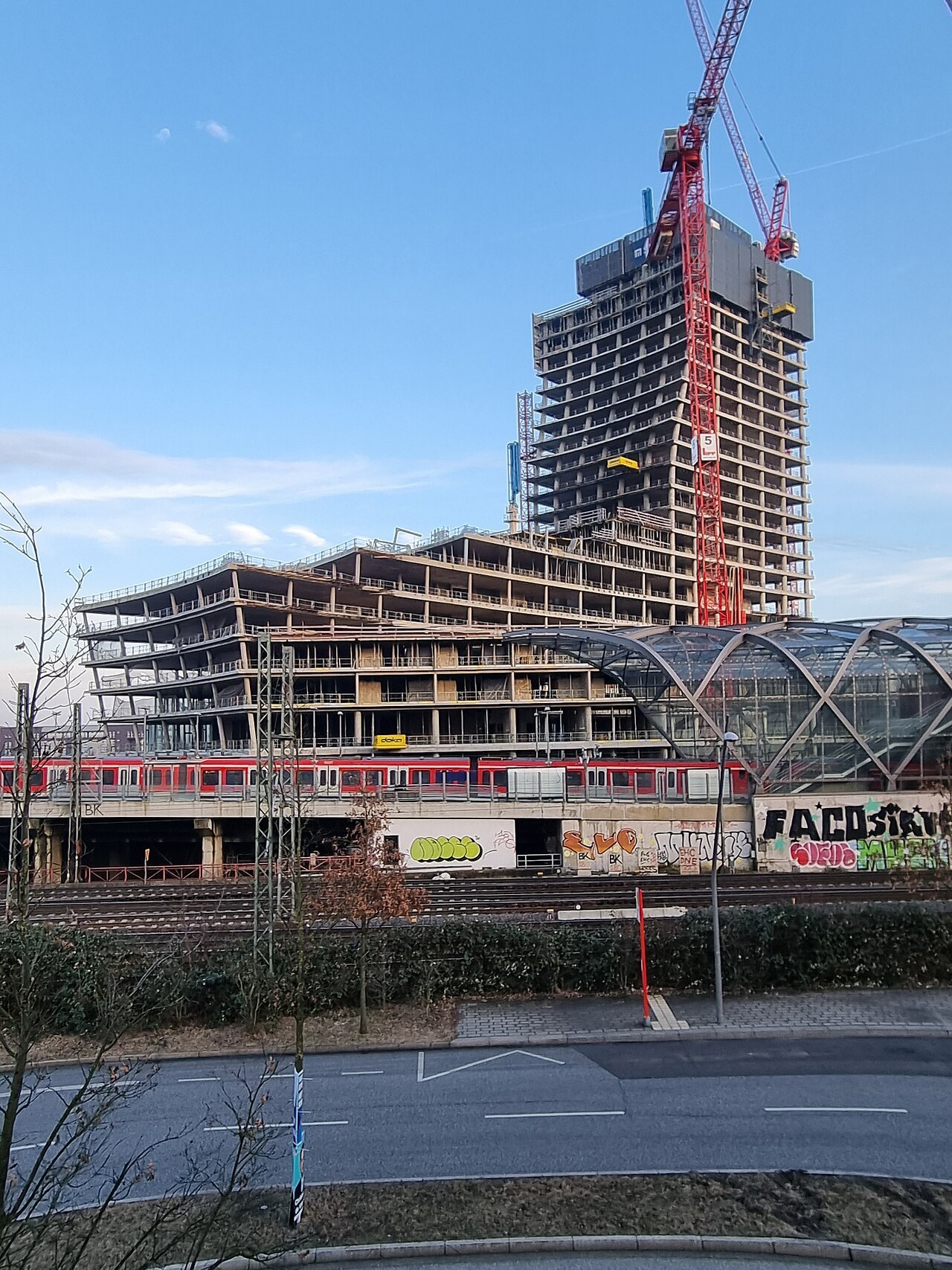 Unfinished concrete frame of the Elbtower in Hamburg with tower cranes, seen from the west in February 2025 beside the glass roof of Elbbrücken station and an S-Bahn train