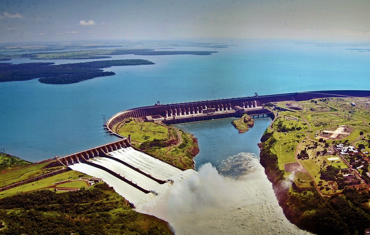 Projects Aerial view of Itaipu Dam on the Paraná River, with the curved main dam and powerhouse behind the spillway discharging into the river channel