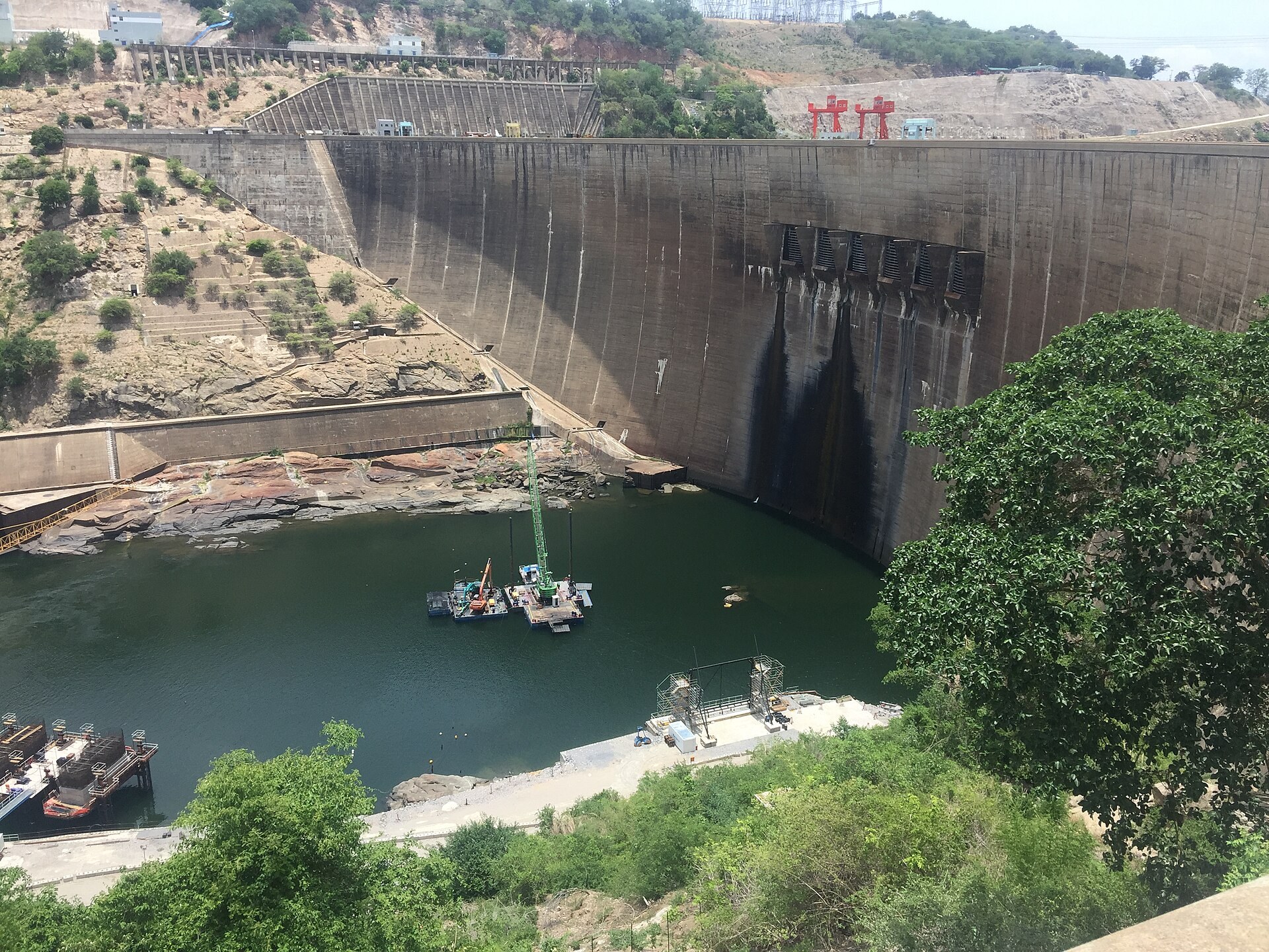 Downstream face of the arch dam with the six spillway openings, a crane barge working in the plunge pool and site works on the bank