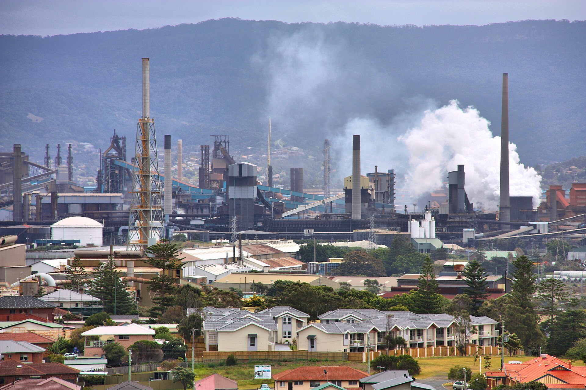 Telephoto of the ironmaking end of Port Kembla Steelworks: blast furnace structures, stoves, stacks and a steam plume, with houses in the foreground and the escarpment behind