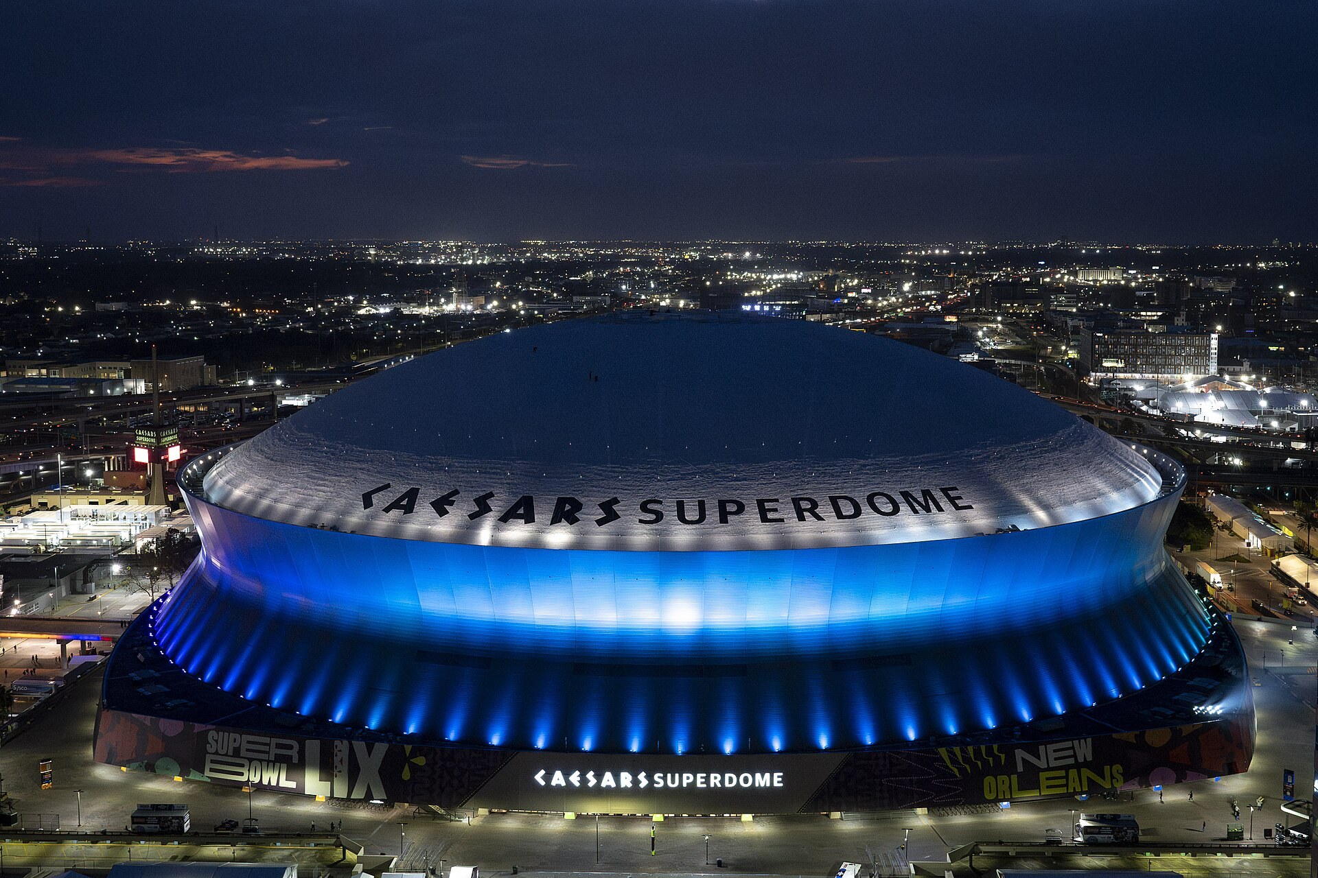 Aerial dusk shot of the full dome lit blue with CAESARS SUPERDOME lettering and Super Bowl LIX graphics at the base